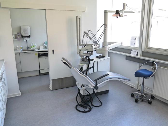 Lyst rum med tandlægestol med udstyr, vinduer og kig til et andet rum med vask og forskellige remedier.