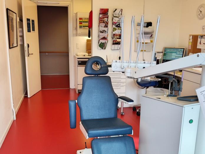 Tandlægestol med forskelligt udstyr, rødt gulv, pjecer og kontorplads i baggrunden.