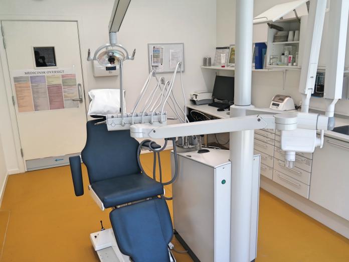 Tandlægestol med forskelligt udstyr og skabe og lille kontorplads i baggrunden.