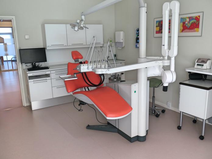 Rød tandlægestol med forskelligt udstyr, skabe og pc-skærm i baggrunden.
