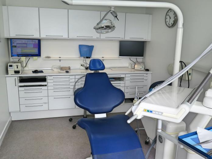 Blå tandlægestol med forskelligt udstyr og lys. Et ur på væggen. Skuffer og skabe i baggrunden og en computerskærm.