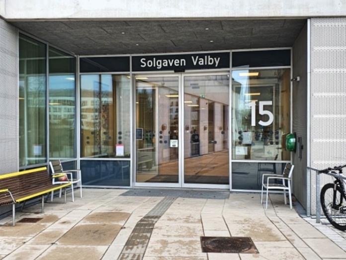 Indgang til Solgaven Valby. Taktile fliser og skydedøre af glas. På dørene står også "Solgaven Valby" og tallet "15". Der er en bænk og et par stole udenfor.