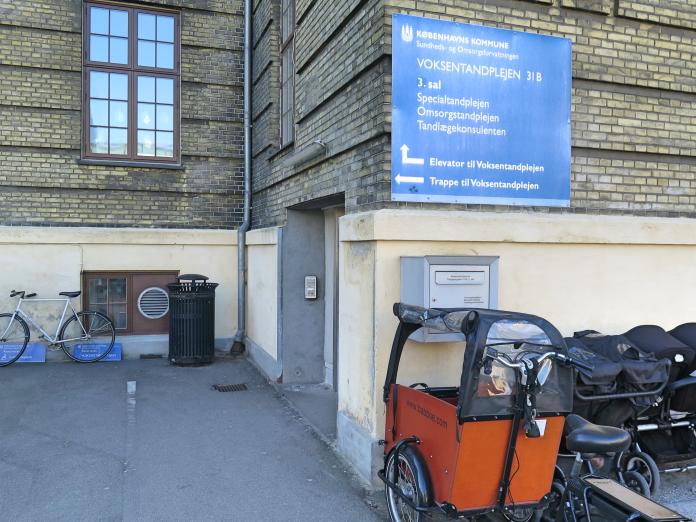 Klinikken fra gaden. Gul murstensbygning og gul sokkel. Blåt skilt med pile til elevator og trappe. 